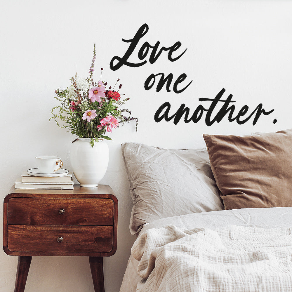 A Love One Another wall decal between a wood nightstand and a bed, available from Tempaper.