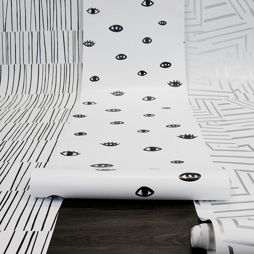 A roll of black and white Eye See You removable wallpaper rolling down the wall and towards us along with other designs from Tempaper.#color_white-and-black-eyes