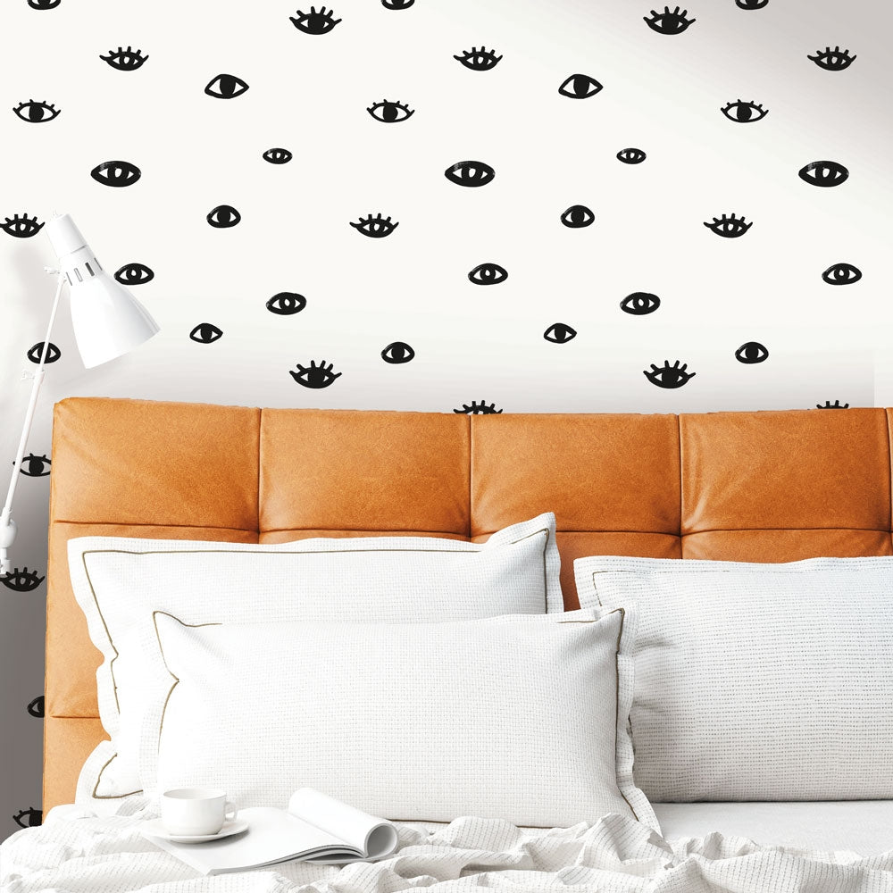 Tempaper's black and white Eye See You removable wallpaper in a bedroom with a leather headboard and white bed sheets.#color_white-and-black-eyes