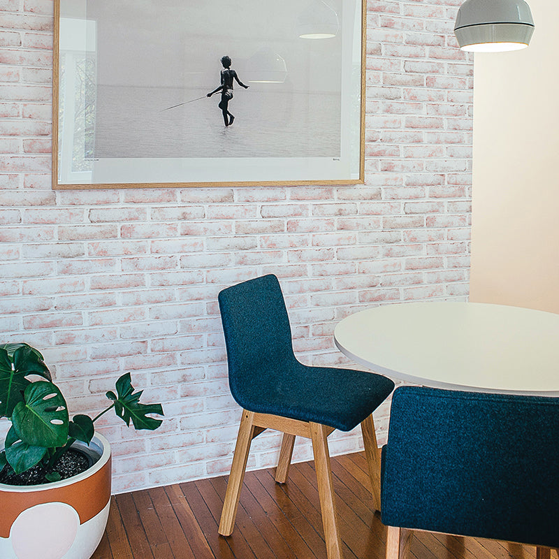 Tempaper's Whitewashed Brick Peel And Stick Wallpaper shown behind a plant and white table with blue chairs.