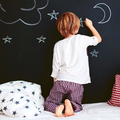 A child kneeling on a bed drawing stars and a moon on chalkboard wallpaper from Tempaper.