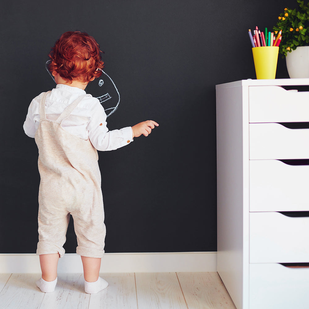 A small toddler drawing a design on Tempaper's chalkboard wallpaper.