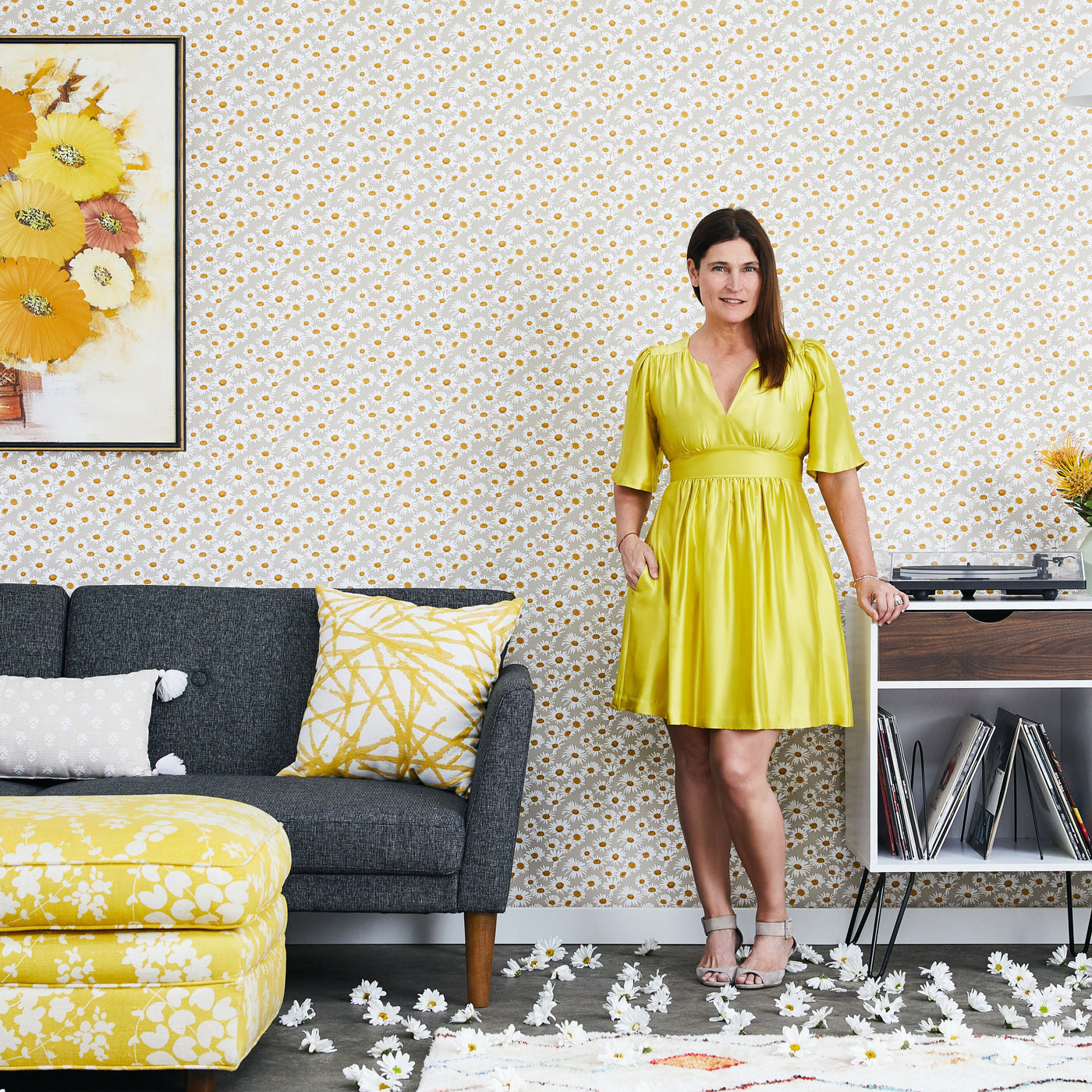 A woman in a yellow dress standing in a living room in front of a wall covered in Tempaper's daisies floral peel-and-stick wallpaper.