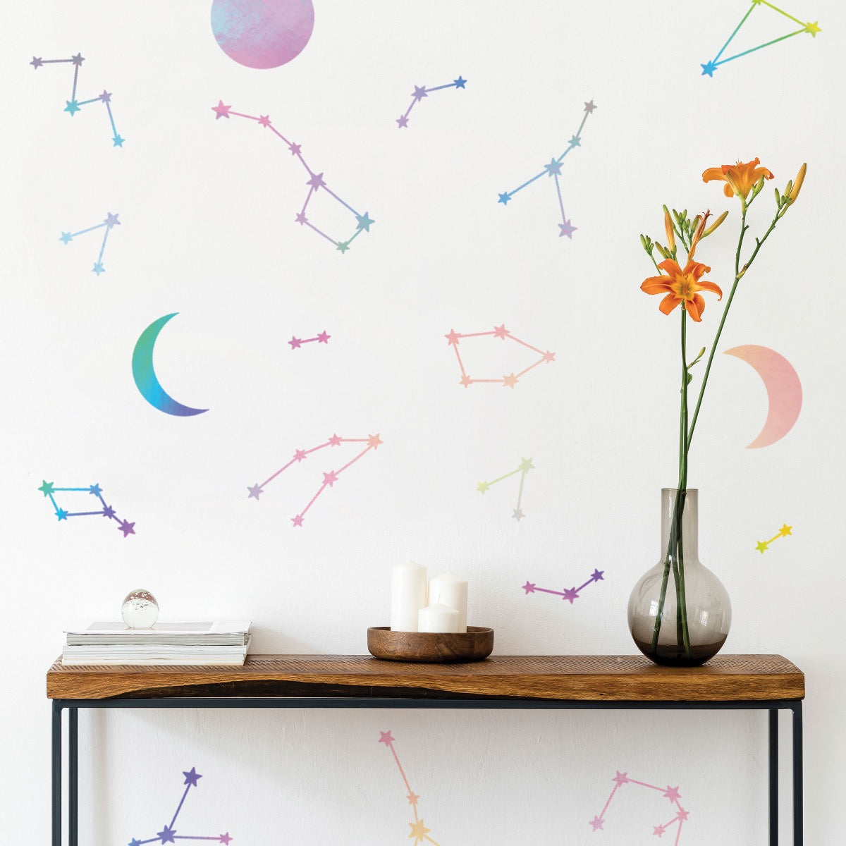 A wood side table with magazines, candles, and flowers in front of a wall designed using Tempaper's holographic Constellations & Moons wall decals.