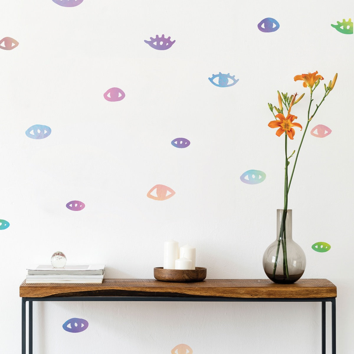 The holographic You Are Seen wall decals on a white wall behind a side table with a candle, flowers, and magazines, available from Tempaper.