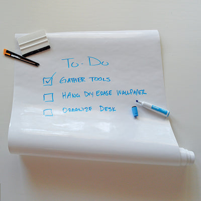 A roll of Tempaper's Dry Erase Peel And Stick Wallpaper shown with a squeegee, utility knife, and blue marker.