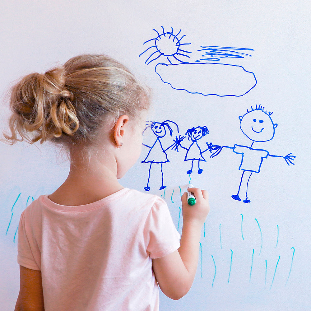 A small child drawing a family and grass on a wall protected with Tempaper's dry erase wallpaper.