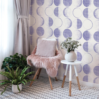 Tempaper's Moons Peel And Stick Wallpaper in a periwinkle colorway behind a white chair and white end table.