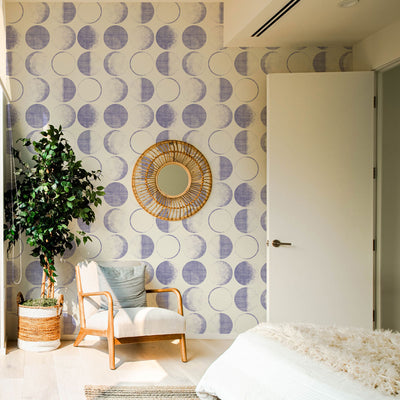 A bedroom with a chair and plant in front of Tempaper's Moons Removable Wallpaper in a periwinkle colorway.