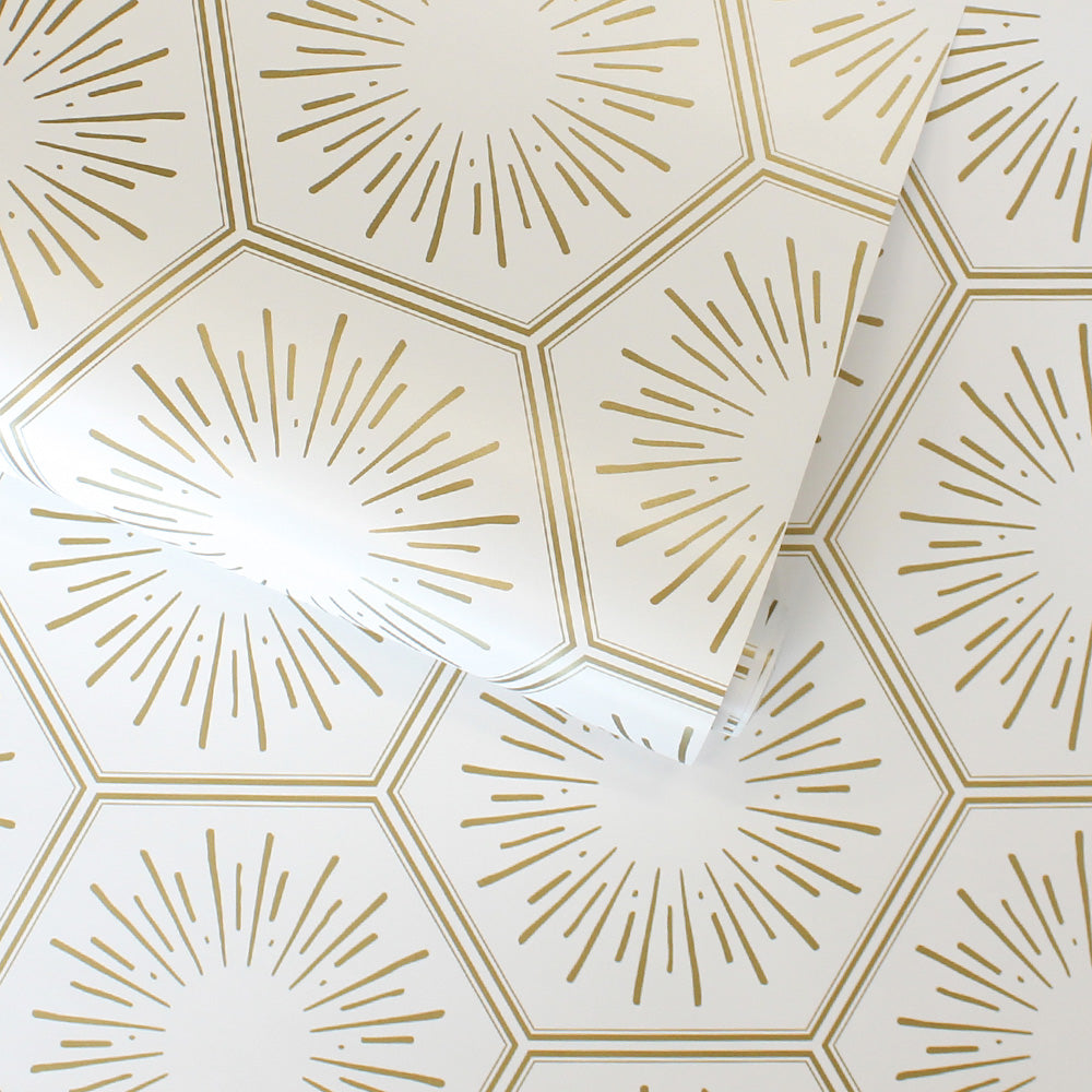A slightly unraveled roll of Tempaper's Hello Sunshine Peel And Stick Wallpaper in metallic gold. Repeating gold hexagons filled with sunbursts — a sample of Tempaper's sunset gold Hello Sunshine peel and stick wallpaper. #color_sunset-gold