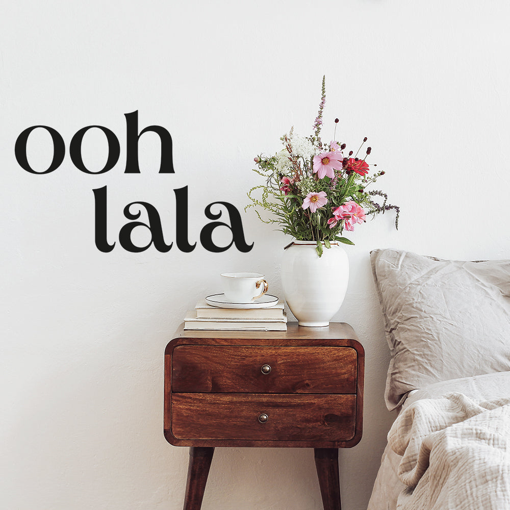 Tempaper's black Ooh La La wall decal on the wall above a wood nightstand decorated with books, a coffee mug, and flowers.