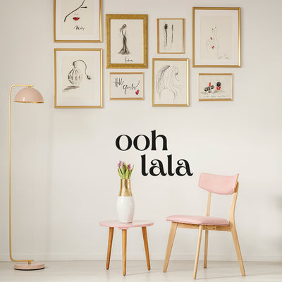A sitting area with a pink chair, table, and lamp with a wall covered in gold frames filled with black sketches and decorated with Tempaper's black Ooh La La wall decals.