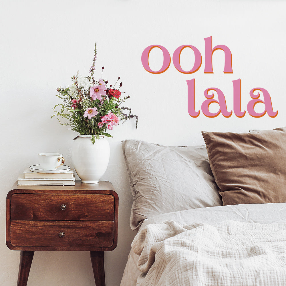 A bedroom with a wood nightstand holding a flower pot, books, and a coffee mug decorated with Tempaper's pink Ooh La La wall decals.