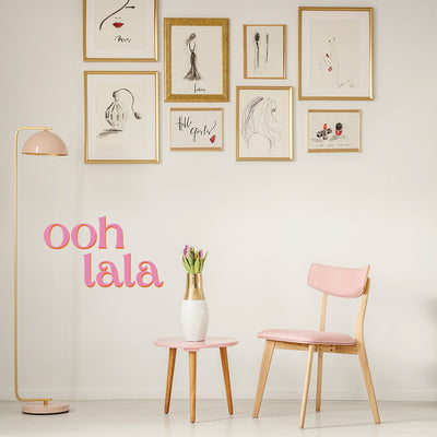 A room with a pink chair and side table and a wall of gold frames filled with black sketches decorated using Tempaper's pink Ooh La La wall decals.