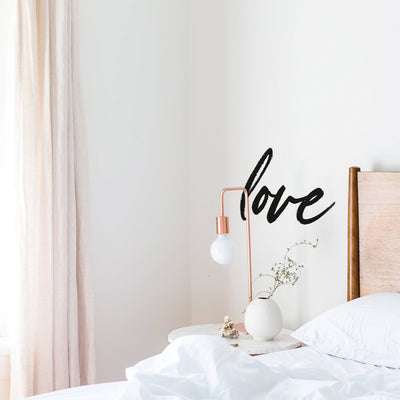 Tempaper's Love wall decal above a white nightstand with a copper lamp and white vase next to a bed.