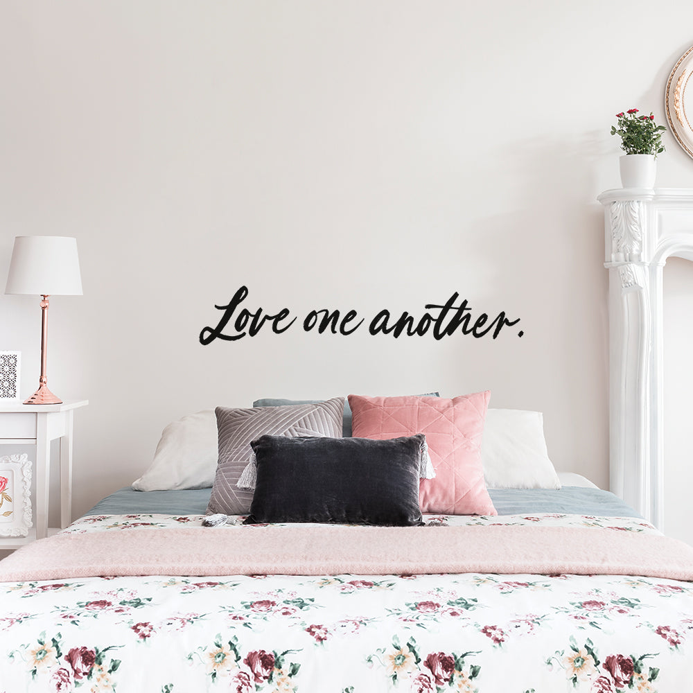Tempaper's Love One Another wall decal on the wall of a bedroom with a white bed and white nightstand.
