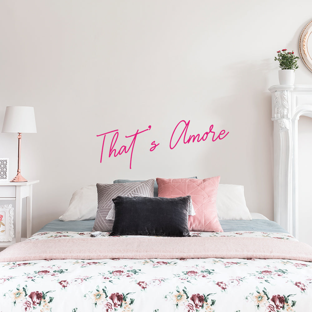 A That's Amore wall decal from Tempaper centered above a bed in a white room with a white nightstand.