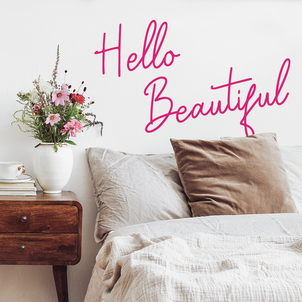 Tempaper's Hello Beautiful wall decal on a wall behind a bed with a wood nightstand holding books, a coffee cup, and a vase of flowers.