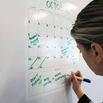 A white woman writing on Tempaper's Dry Erase Monthly Calendar Wall Decal.
