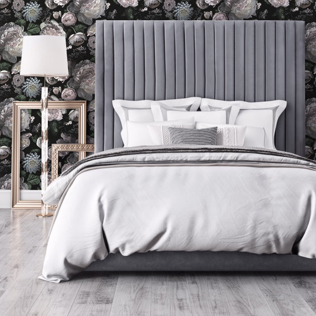A bed with a floor lamp, framed propped against the wall, a tall grey fabric headboard, and Moody floral peel-and-stick wallpaper from Tempaper on the wall.