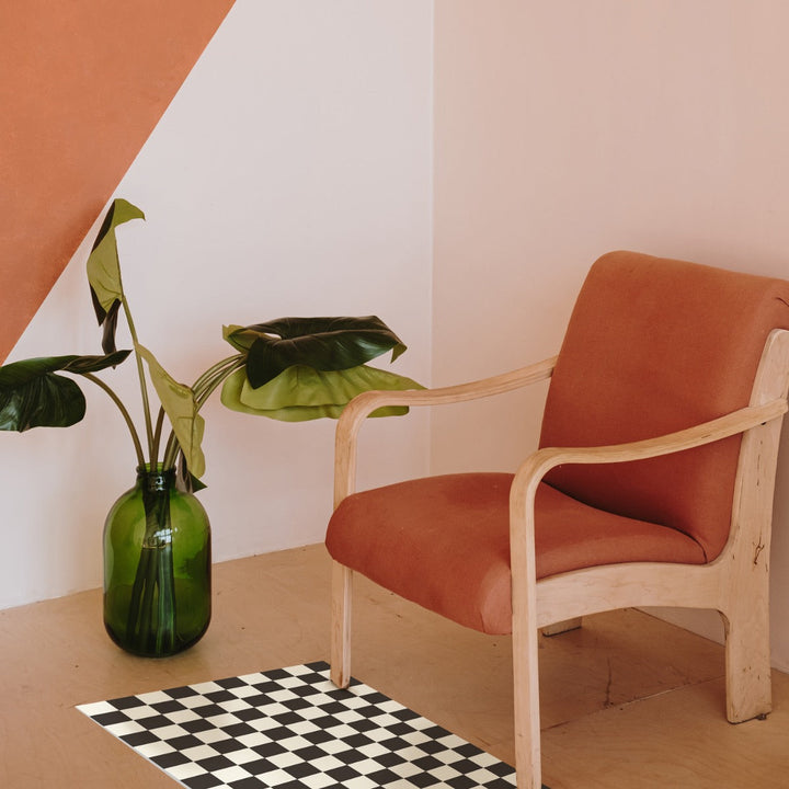 Tempaper's Checkmate Vinyl Rug in black and white shown under a chair and plant.#color_domino