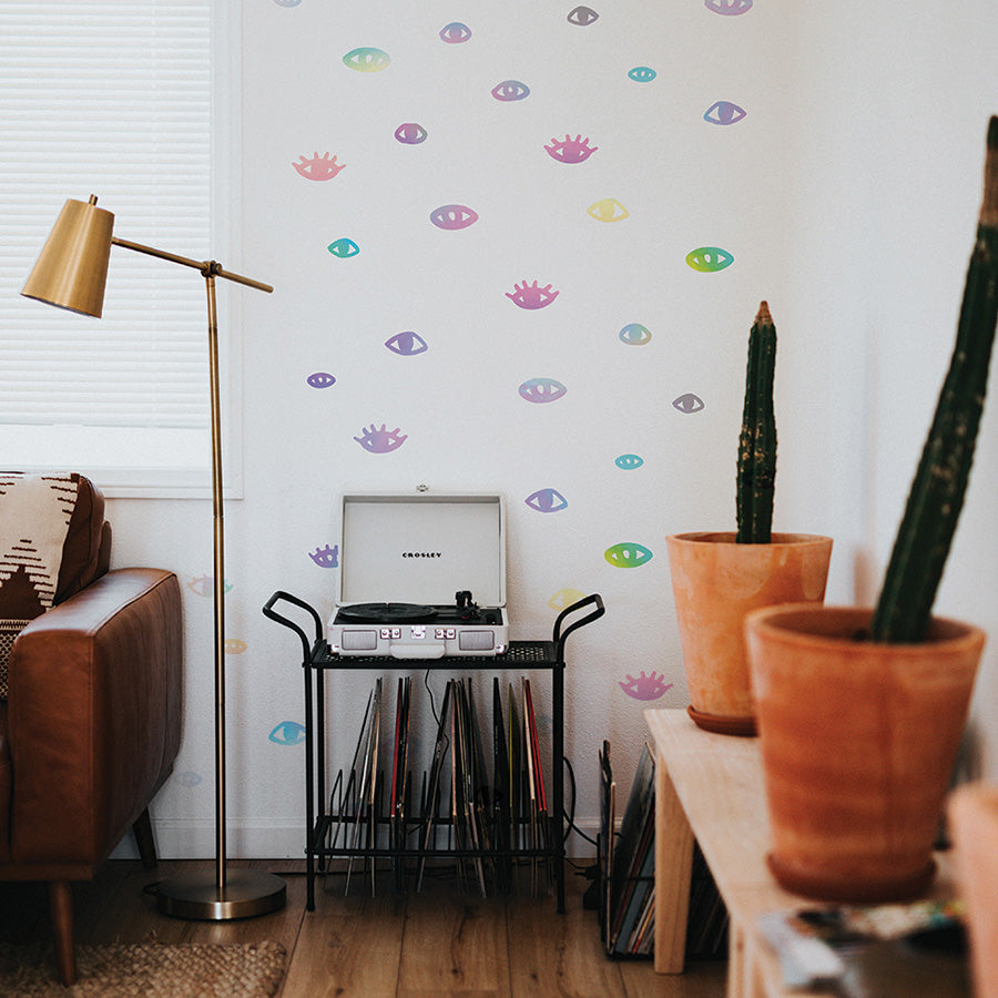 Tempaper's holographic You Are Seen wall decals on a white wall in a living room with a record player and a table with plants.